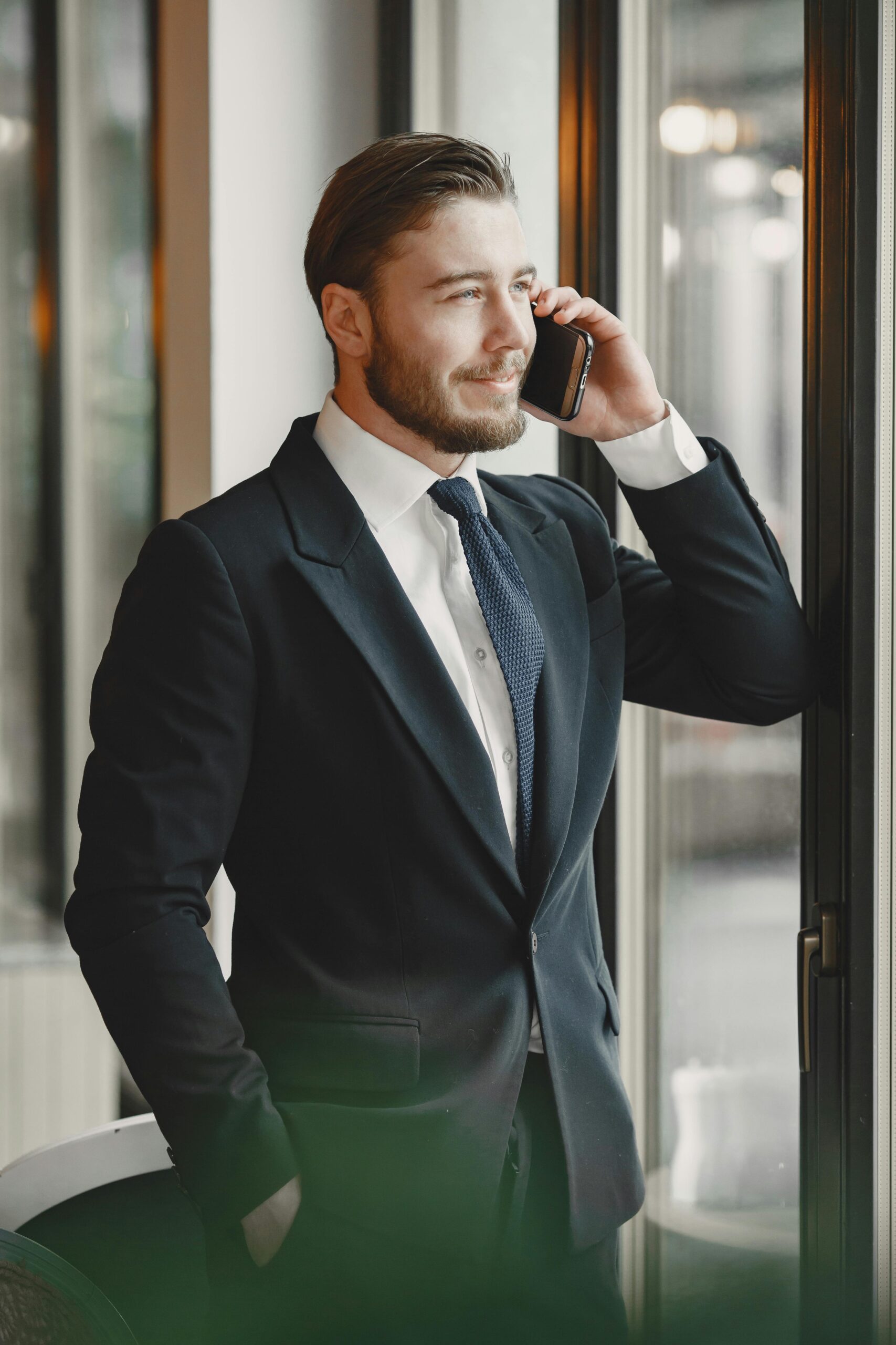 A businessman in a suit talks on the phone by a window, exuding confidence and professionalism.